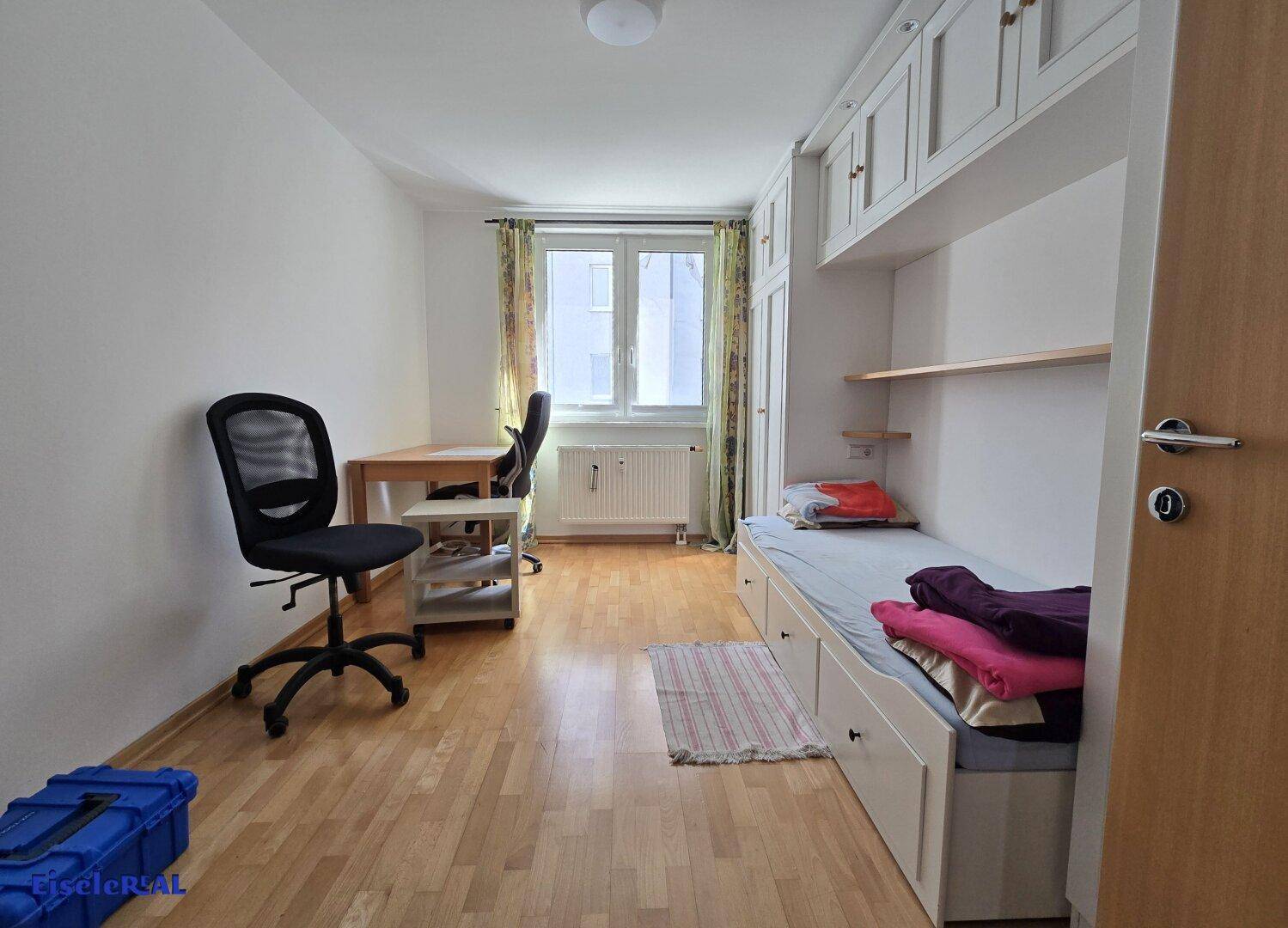 Helles Zimmer mit Bett, Schreibtisch, Parkettboden und Einbauschränken.