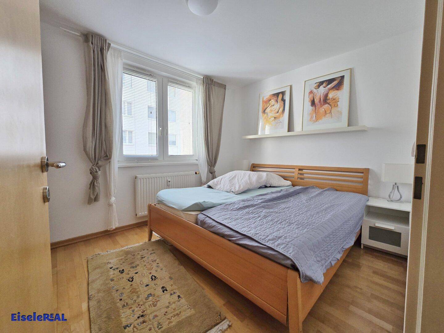 Schlafzimmer mit Doppelbett, Parkettboden und einem Fenster mit Vorhängen.
