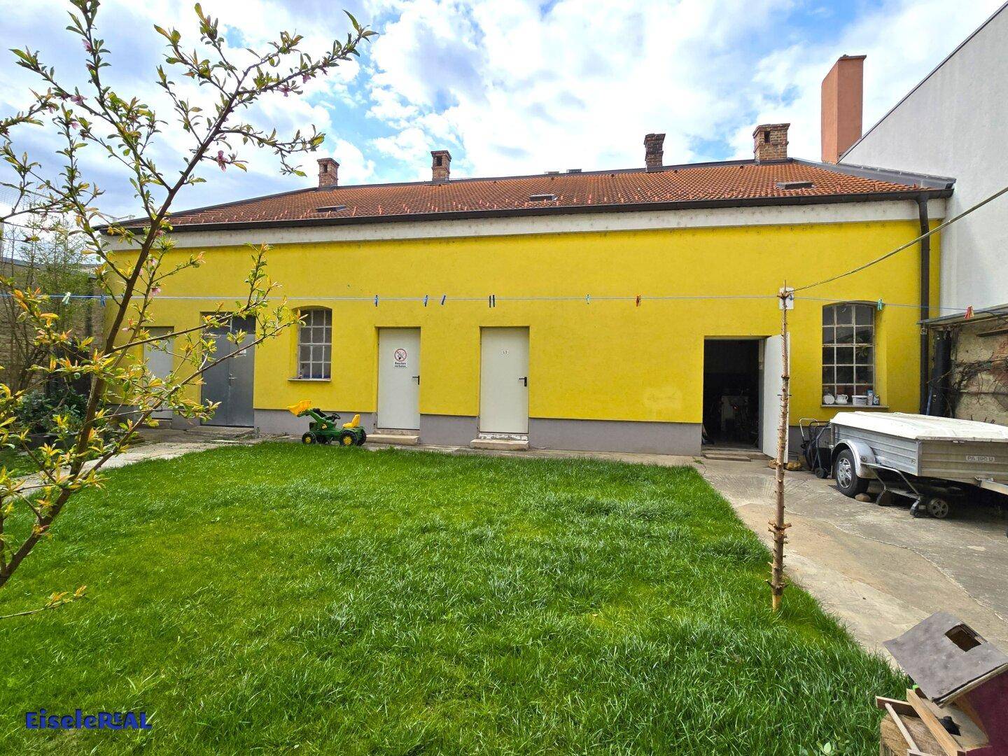 Weitläufiger Rasenbereich im Hinterhof mit einem gelben Nebengebäude und einem Anhänger.