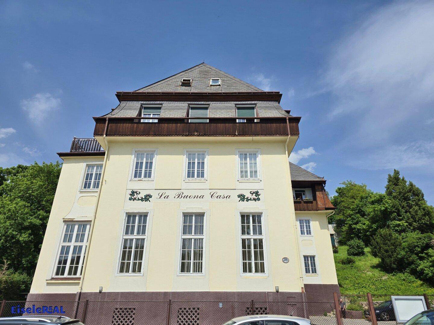 Ansicht des historischen Gebäudes mit gelber Fassade, Mansarddach und vielen Fenstern, umgeben von Grün.