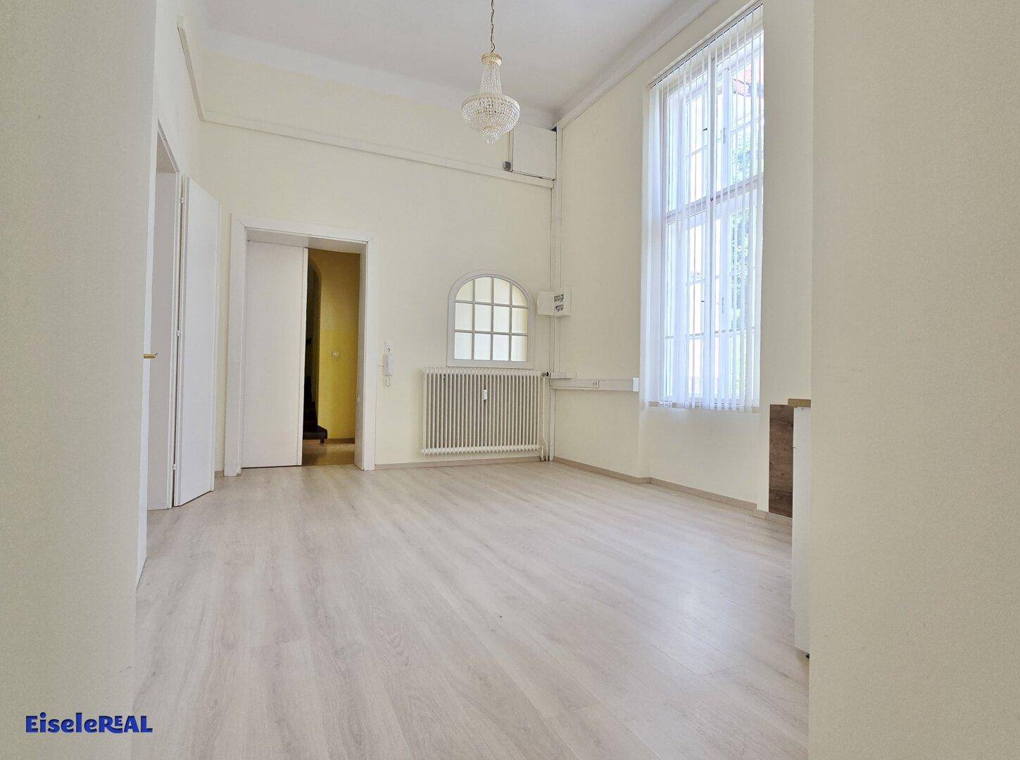 Geräumiger, heller Raum mit hohen Decken, Laminatboden und einem großen, hohen Fenster mit viel Tageslicht.