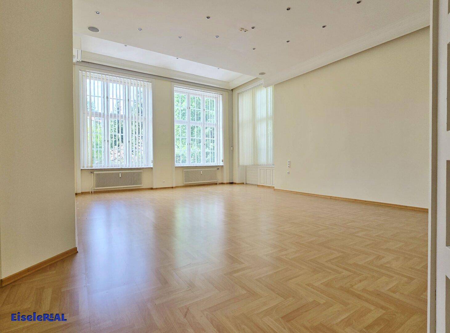 Großer Raum mit Fischgrätparkett, hohen Decken und vielen Fenstern, die den Raum mit Licht durchfluten.