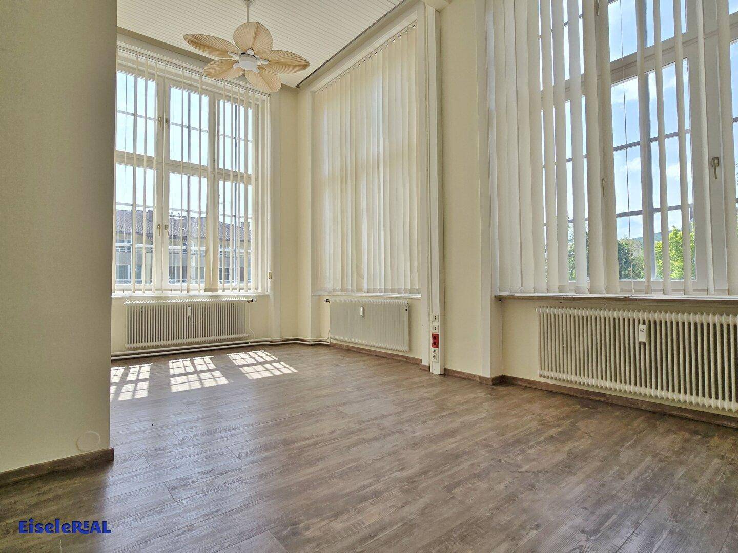 Heller Raum mit Holzoptik-Boden, hohen Fenstern und einem Deckenventilator, der für Belüftung sorgt.