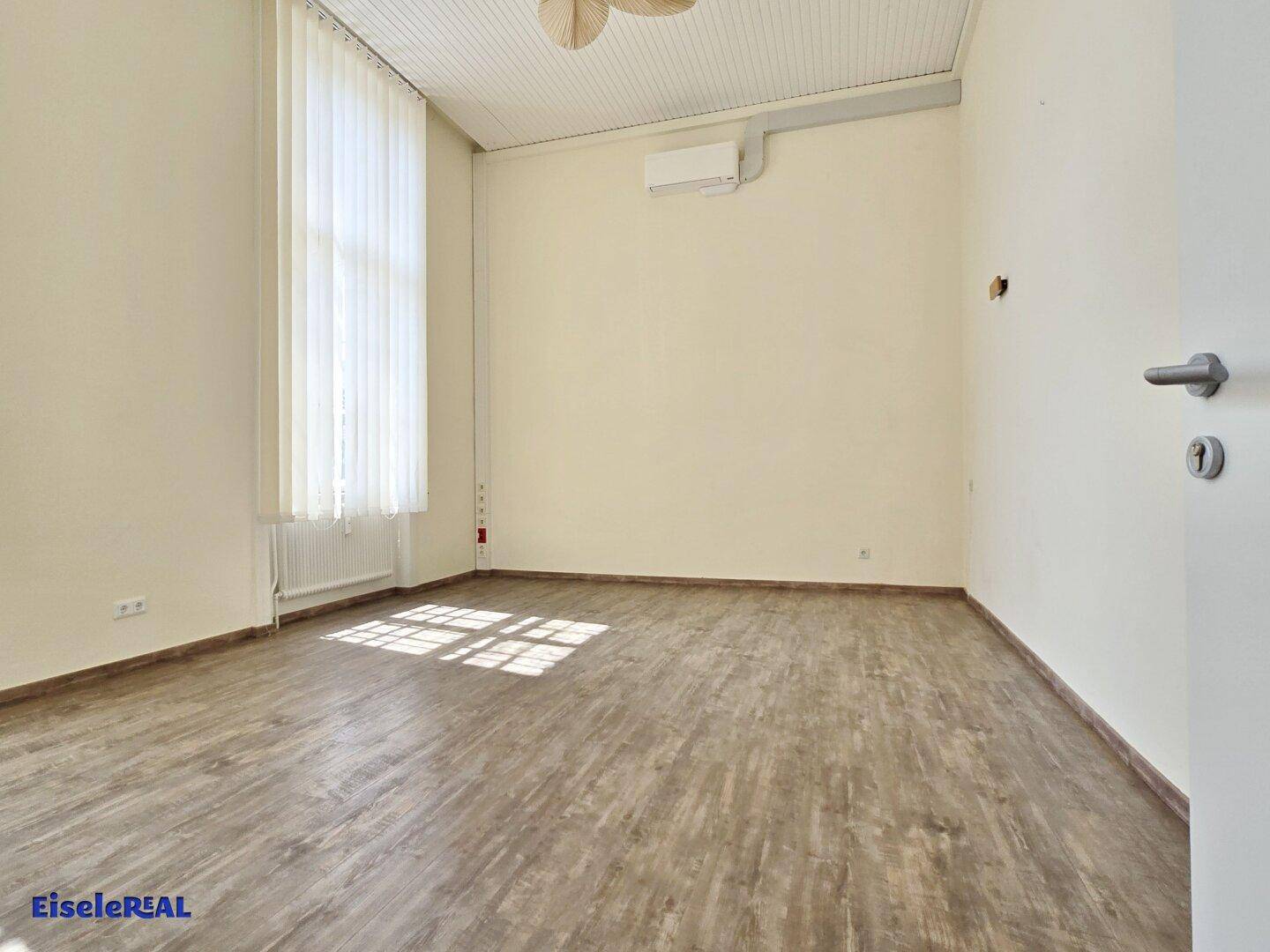Heller Raum mit Holzoptik-Boden, hohen Decken und einem einzelnen hohen Fenster, das Tageslicht hereinlässt.