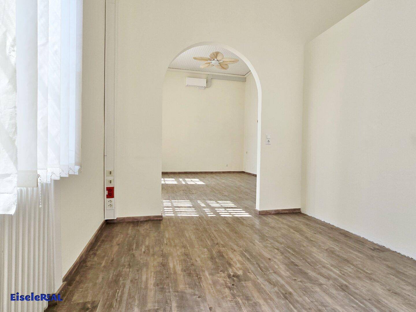 Heller Raum mit Holzoptik-Boden, hohen Decken und einem Bogen, der in einen weiteren, lichtdurchfluteten Bereich führt.