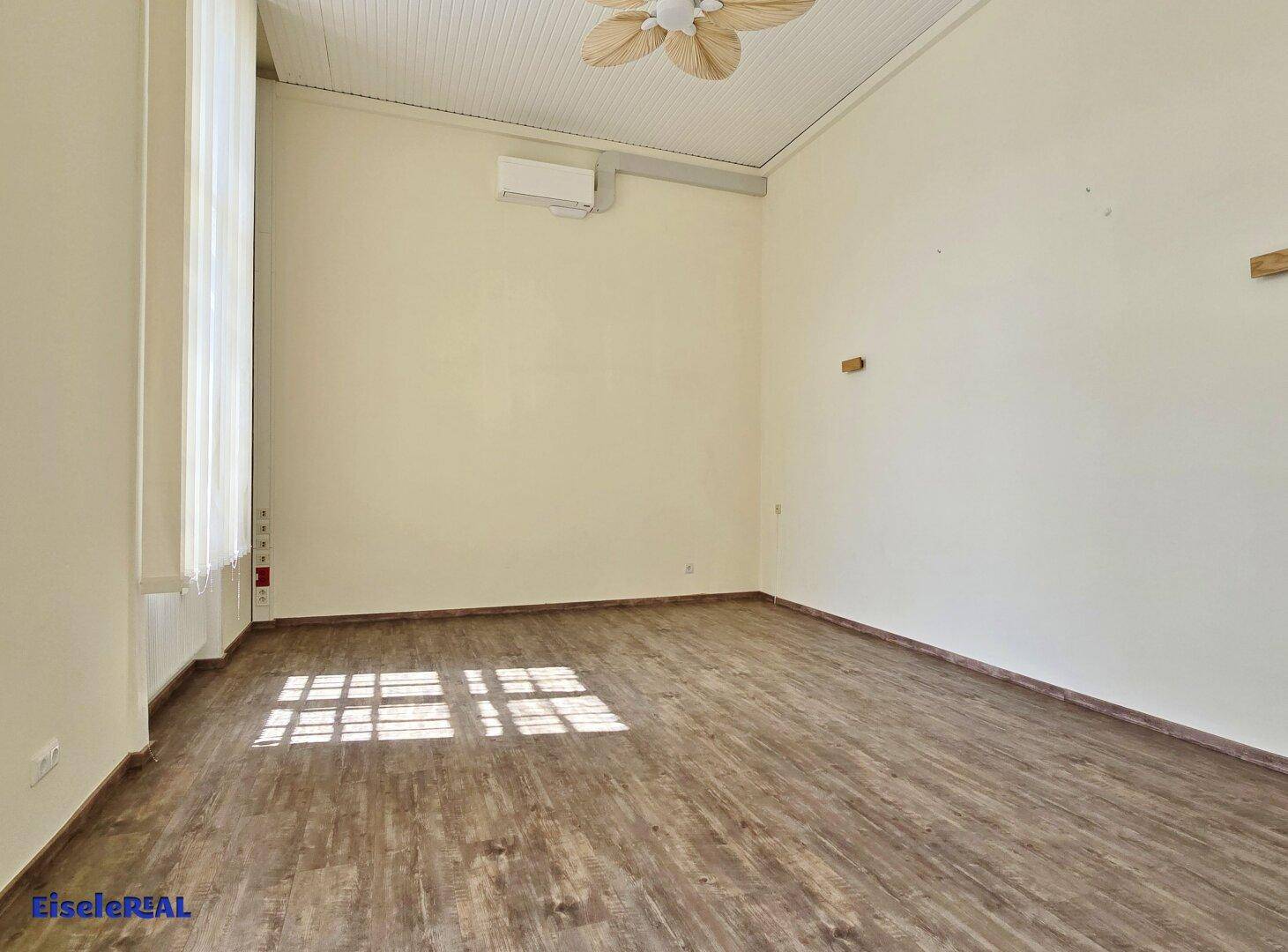 Heller Raum mit Holzoptik-Boden, hohen Decken und einem hohen Fenster, das viel natürliches Licht spendet.