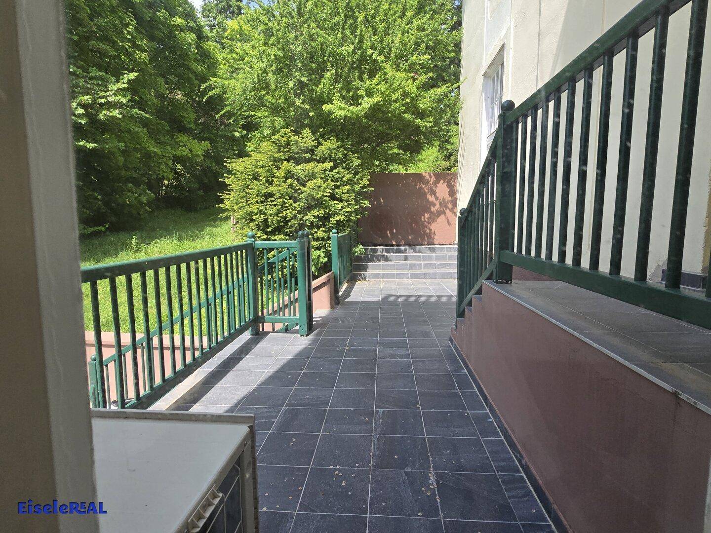 Geflieste Terrasse mit grünem Metallgeländer und einem schönen Blick ins Grüne und auf Bäume.