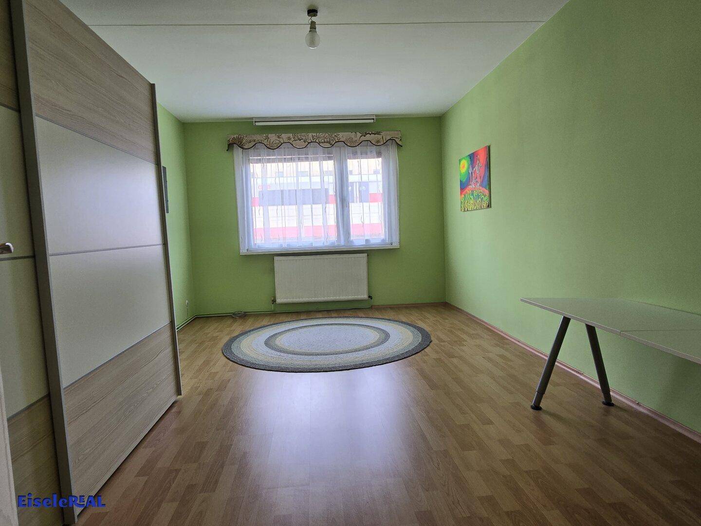 Leeres Zimmer mit hellgrünen Wänden, Holzboden und großem Fenster.