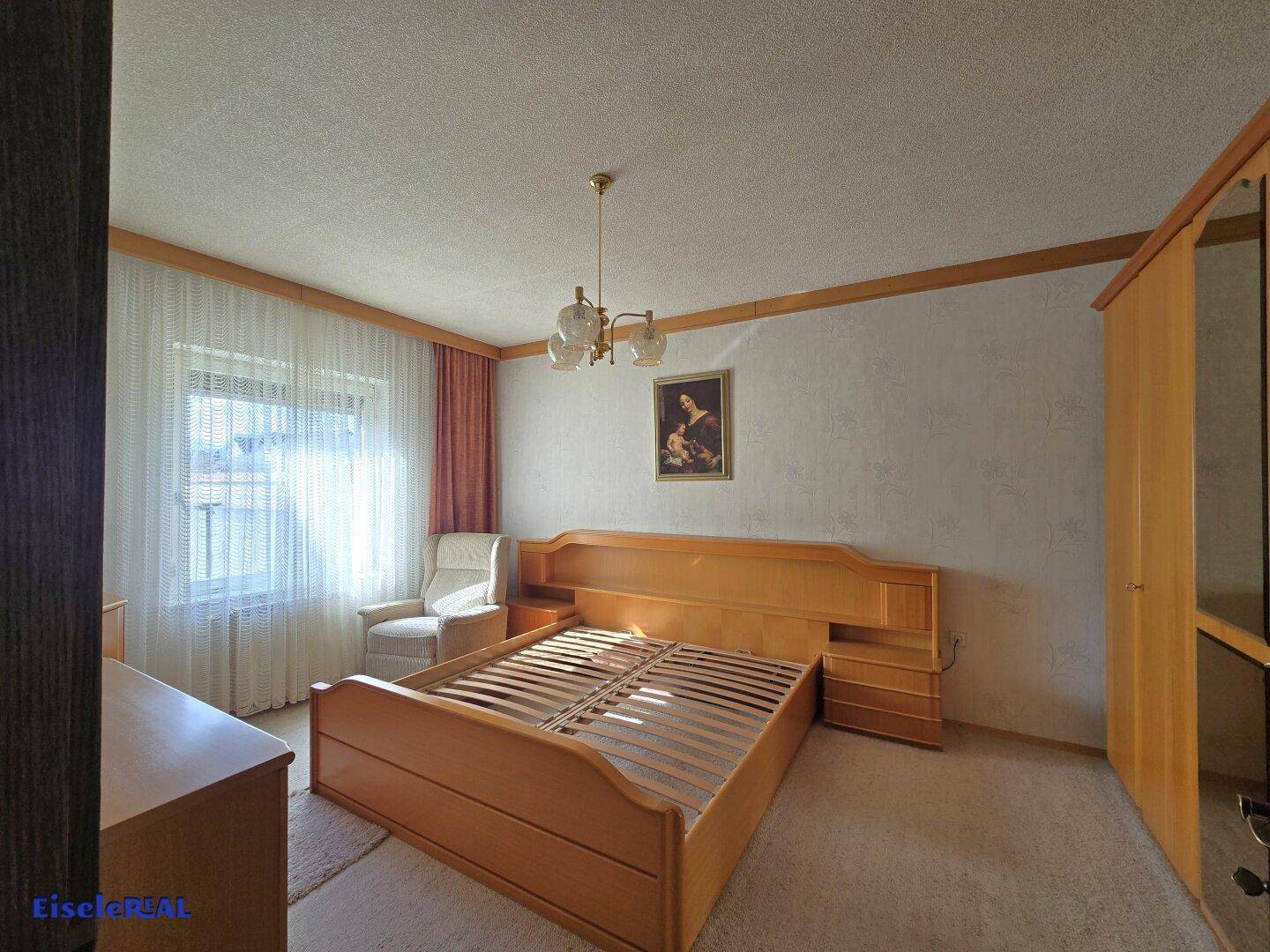 Schlafzimmer mit hellem Holzbett, Teppichboden und großem Fenster.