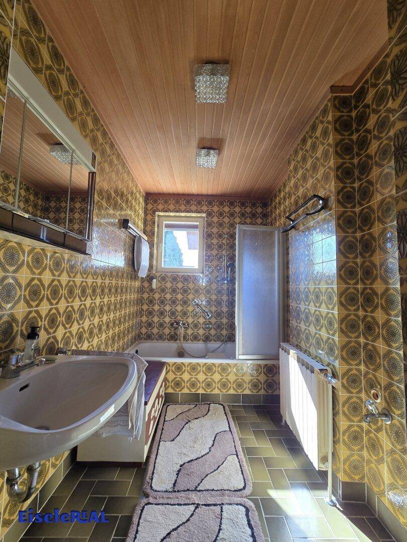 Badezimmer im 80er-Jahre-Stil mit gemusterten Fliesen, Badewanne und Holzdecke.