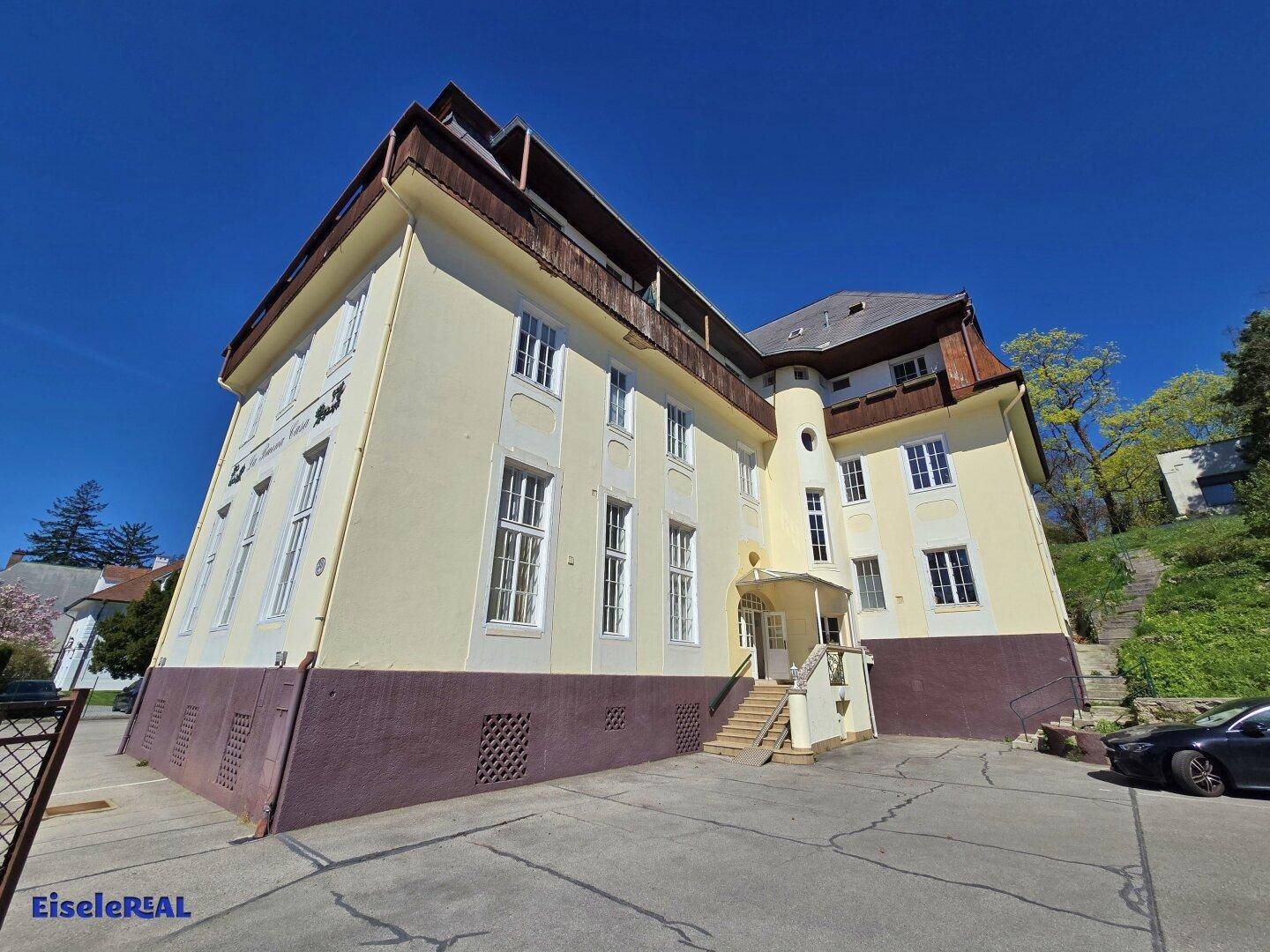 Historisches Gebäude mit gelber Fassade, Mansarddach und angrenzendem Parkplatz.