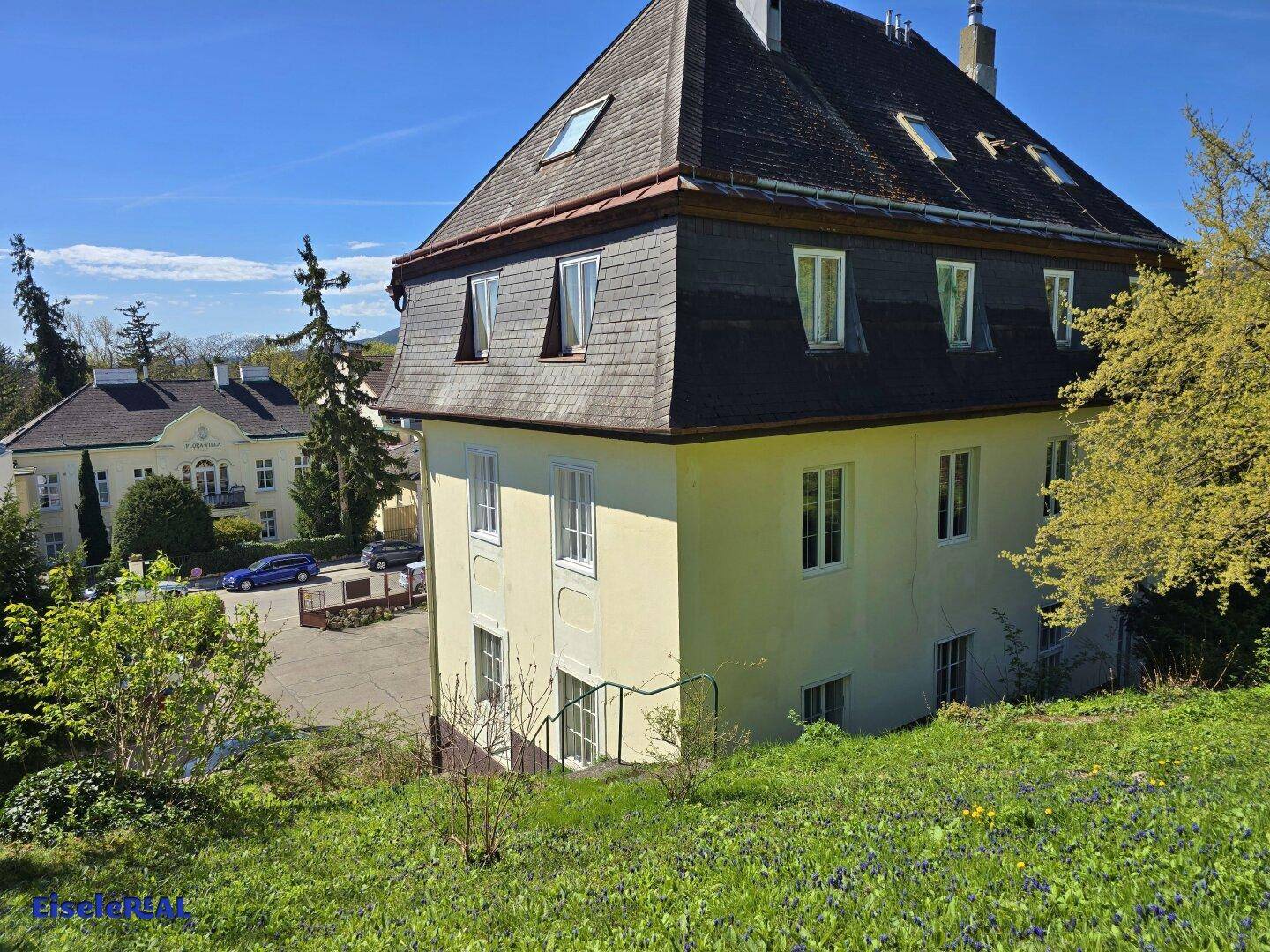 Historisches Gebäude mit Mansarddach, umgeben von grüner Landschaft und Bäumen.