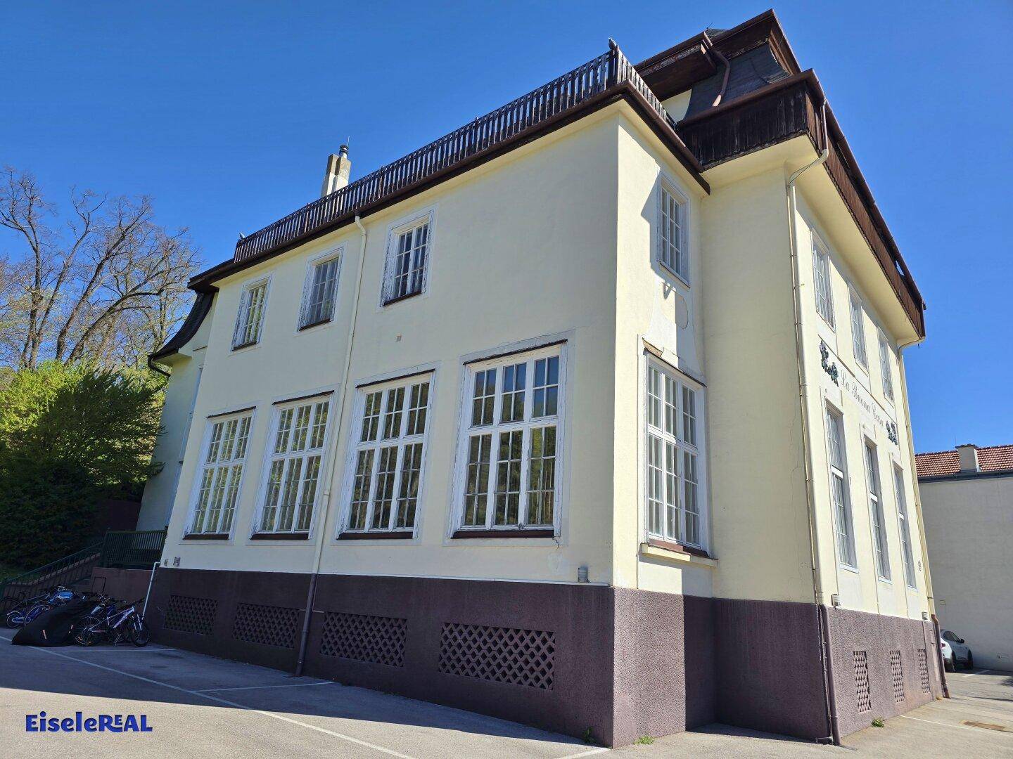 Historisches Gebäude mit gelber Fassade, Mansarddach und angrenzendem Parkplatz.