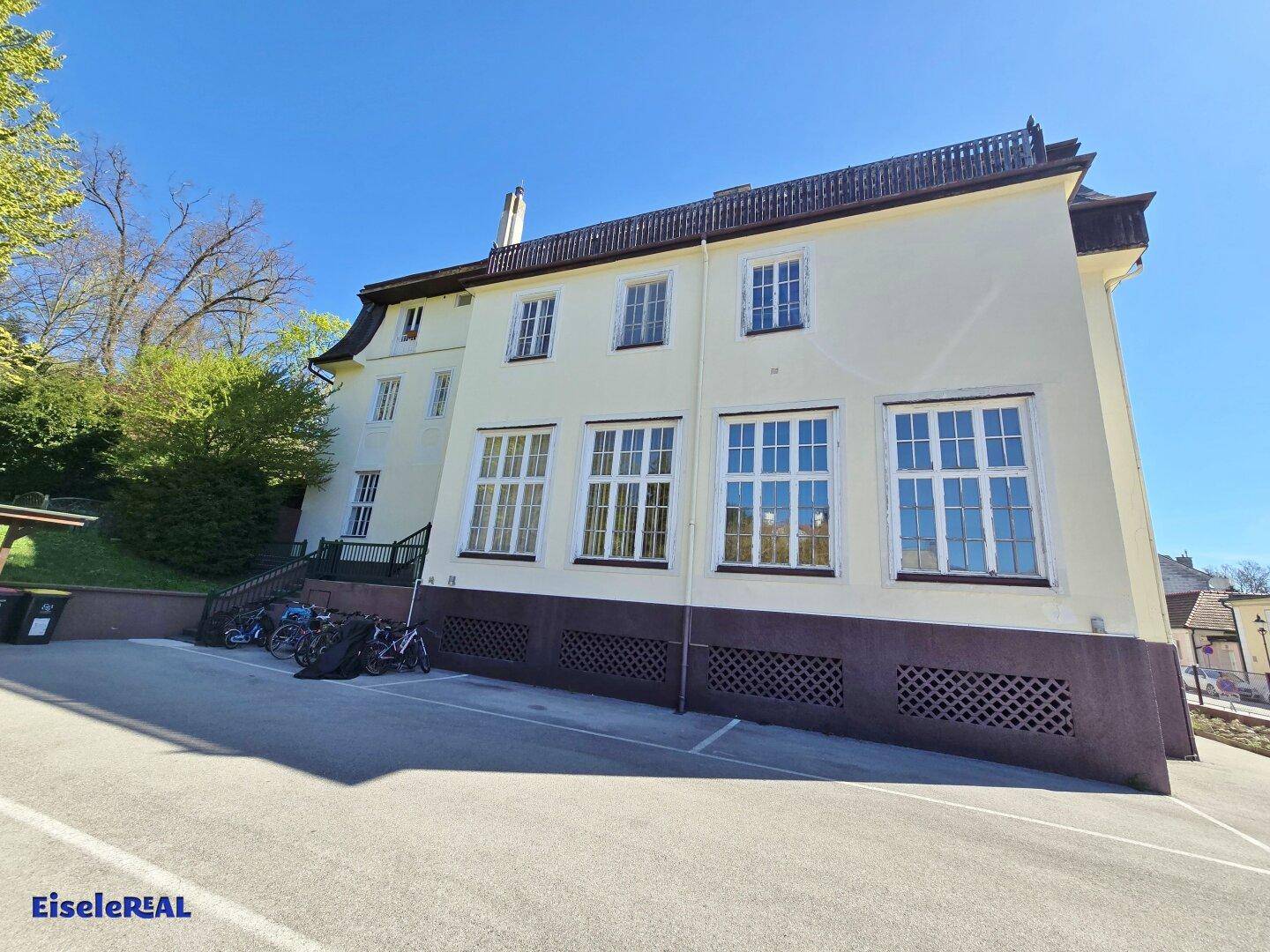 Historisches Gebäude mit gelber Fassade, Mansarddach und angrenzendem Parkplatz.
