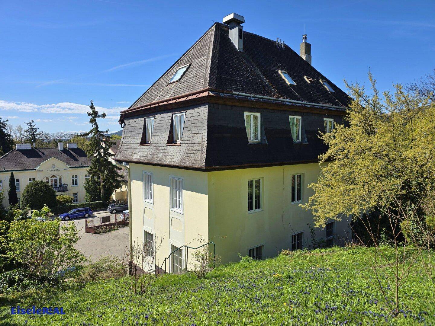 Historisches Gebäude mit Mansarddach, umgeben von grüner Landschaft und Bäumen.