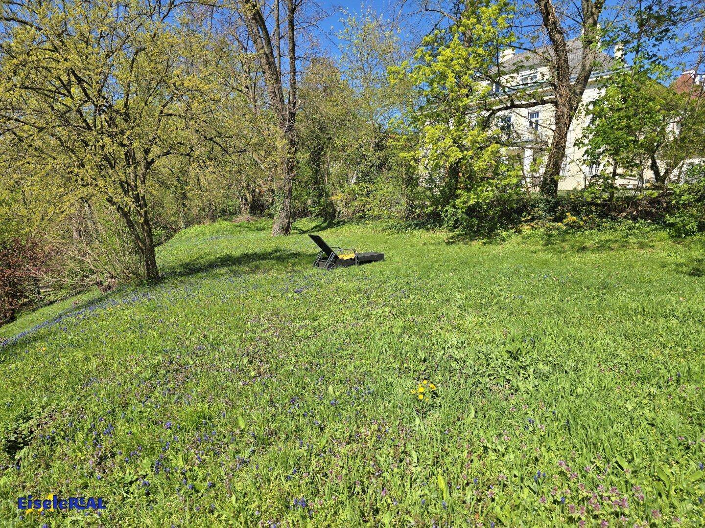 Sonniger Gartenbereich mit Liegestuhl auf einer grünen Wiese unter Bäumen.