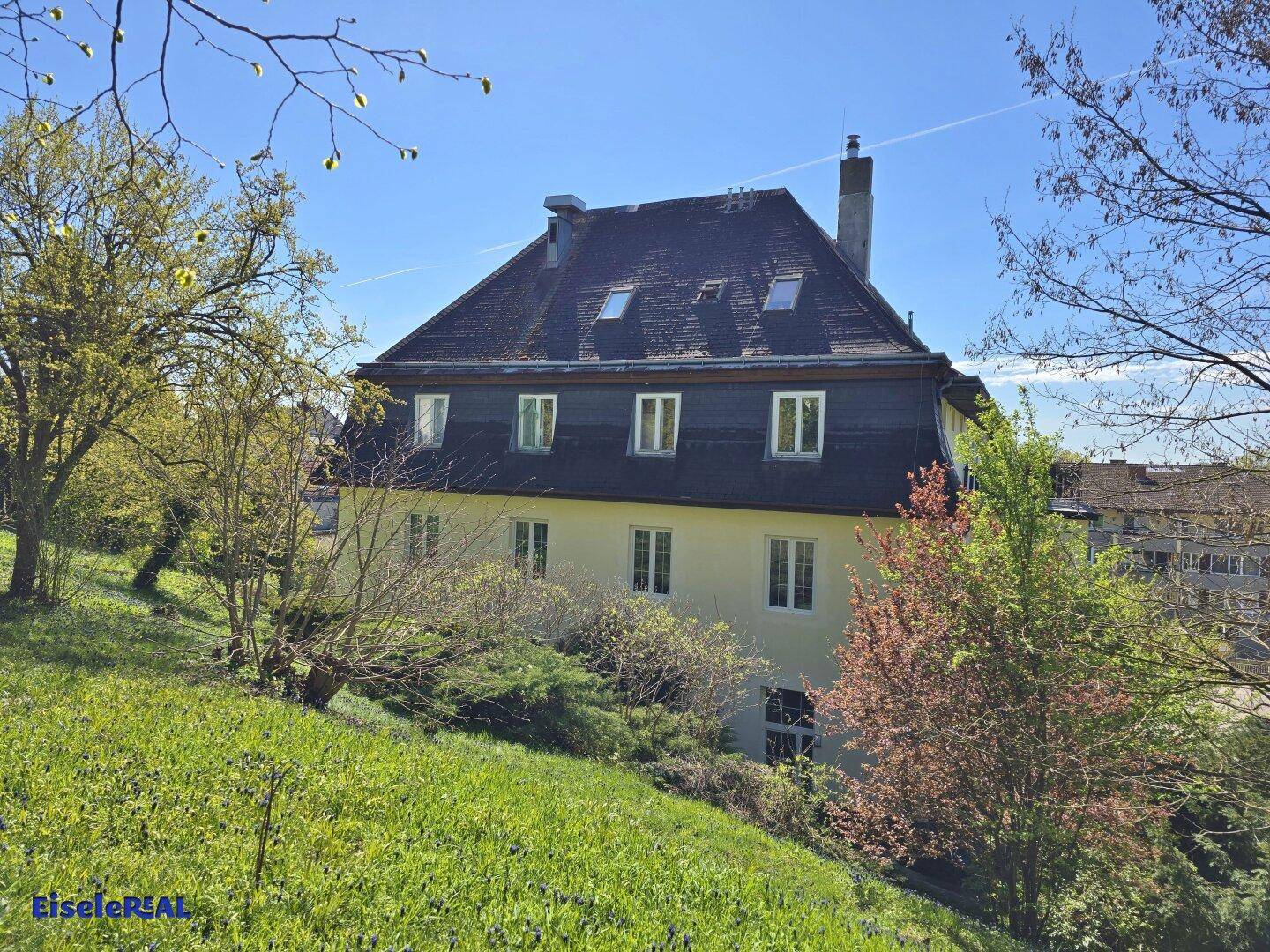 Historisches Gebäude mit Mansarddach, umgeben von grüner Landschaft und Bäumen.
