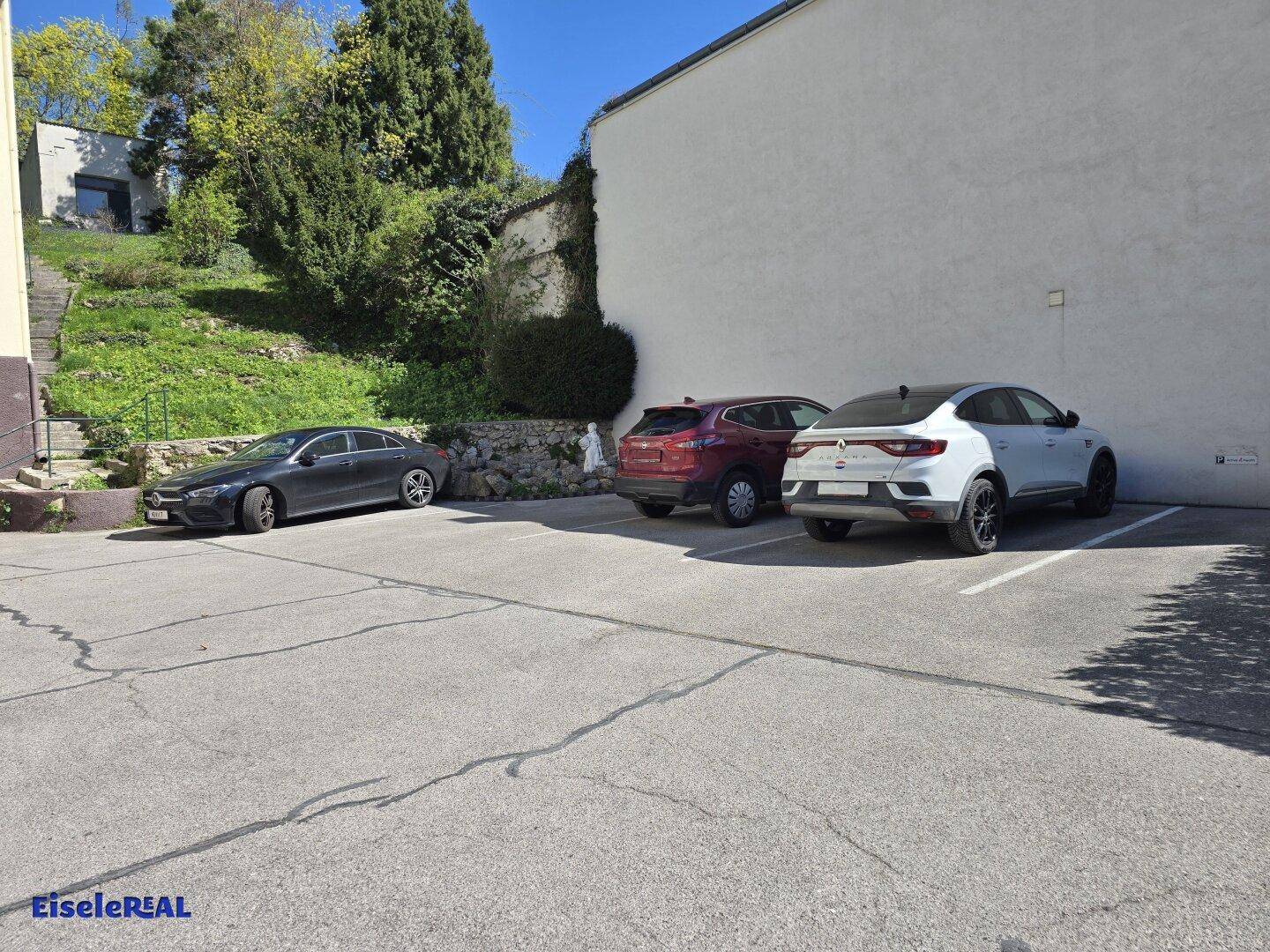 Außenparkplatz mit mehreren geparkten Autos und einer grünen Böschung im Hintergrund.