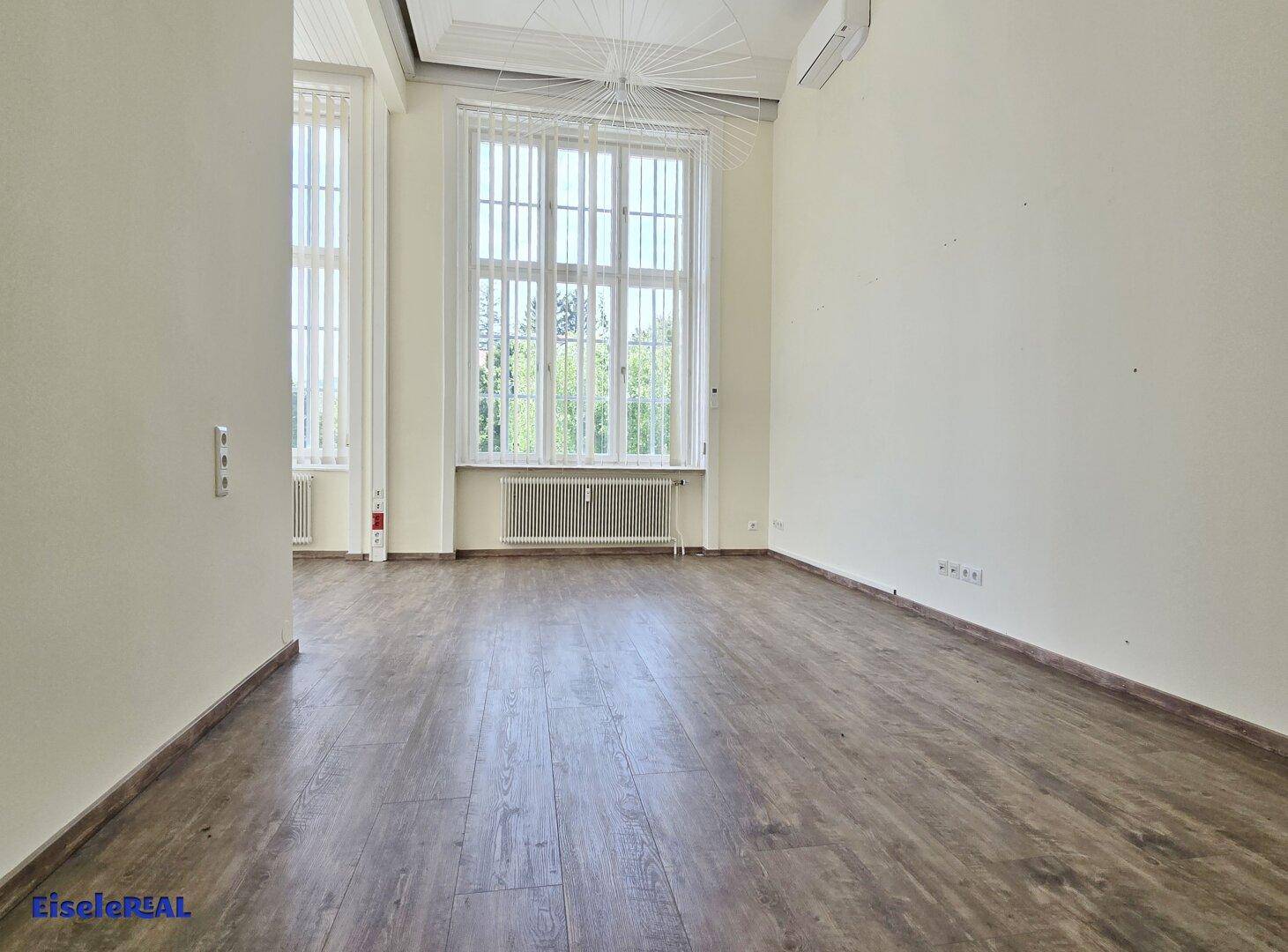 Heller Raum mit Laminatboden und hohen Fenstern, die viel Tageslicht hereinlassen.
