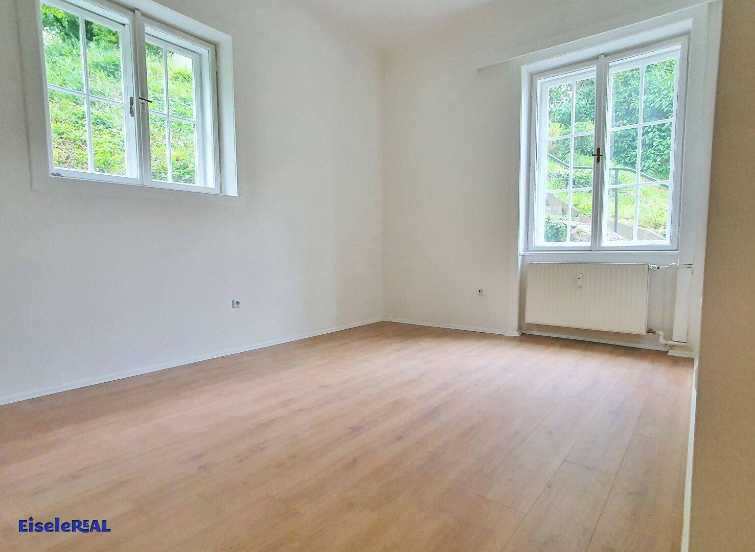 Helles Zimmer mit Holzboden und zwei großen Fenstern mit Blick ins Grüne.