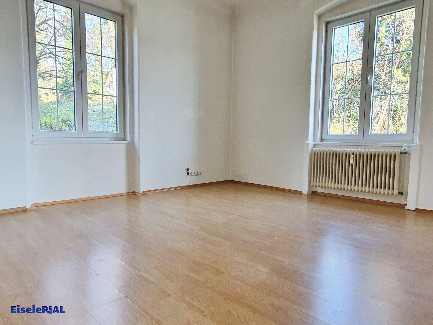 Helles Zimmer mit Holzboden und zwei großen Fenstern, die viel Licht hereinlassen.