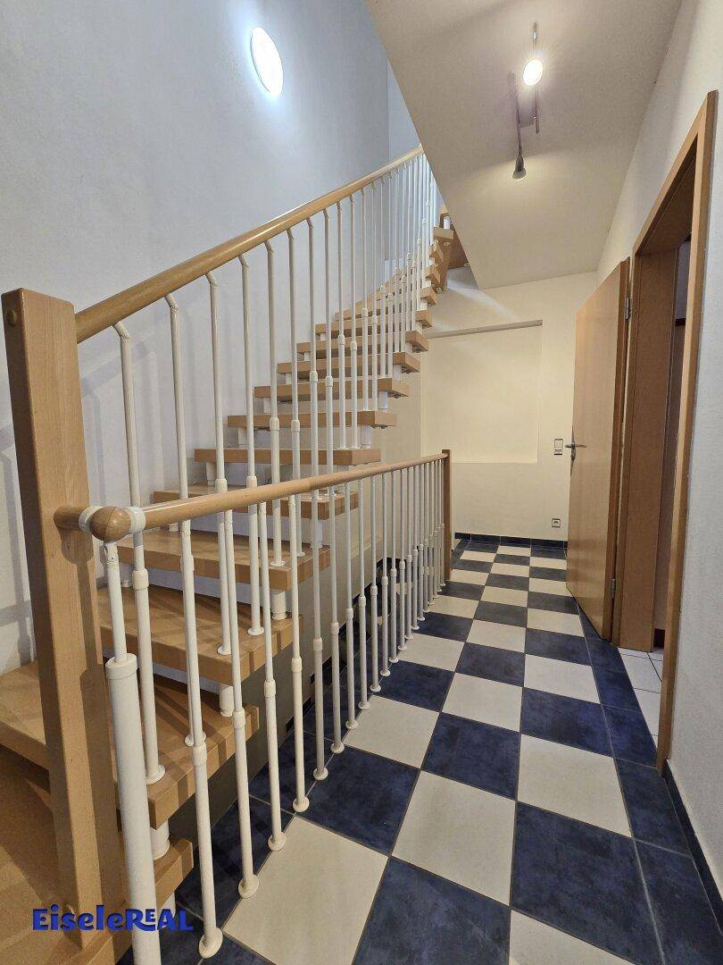 Heller Flur mit Holztreppe und blau-weiß kariertem Fliesenboden, der zu den oberen Etagen führt.
