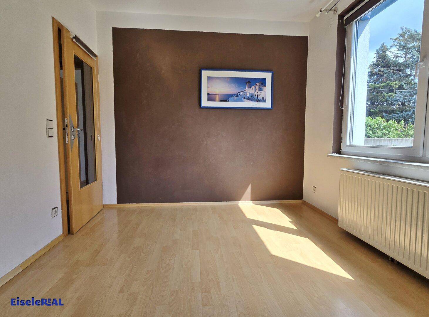 Leerer Raum mit hellem Laminatboden, einer braunen Akzentwand und einem großen Fenster.