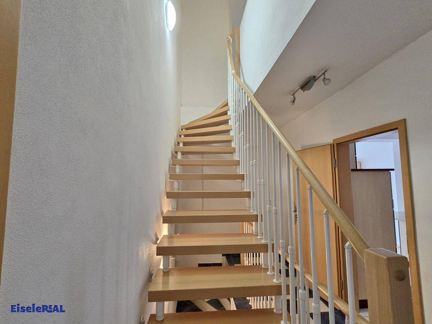 Helle Holztreppe mit weißem Geländer, die in die obere Etage führt, beleuchtet durch ein Oberlicht.