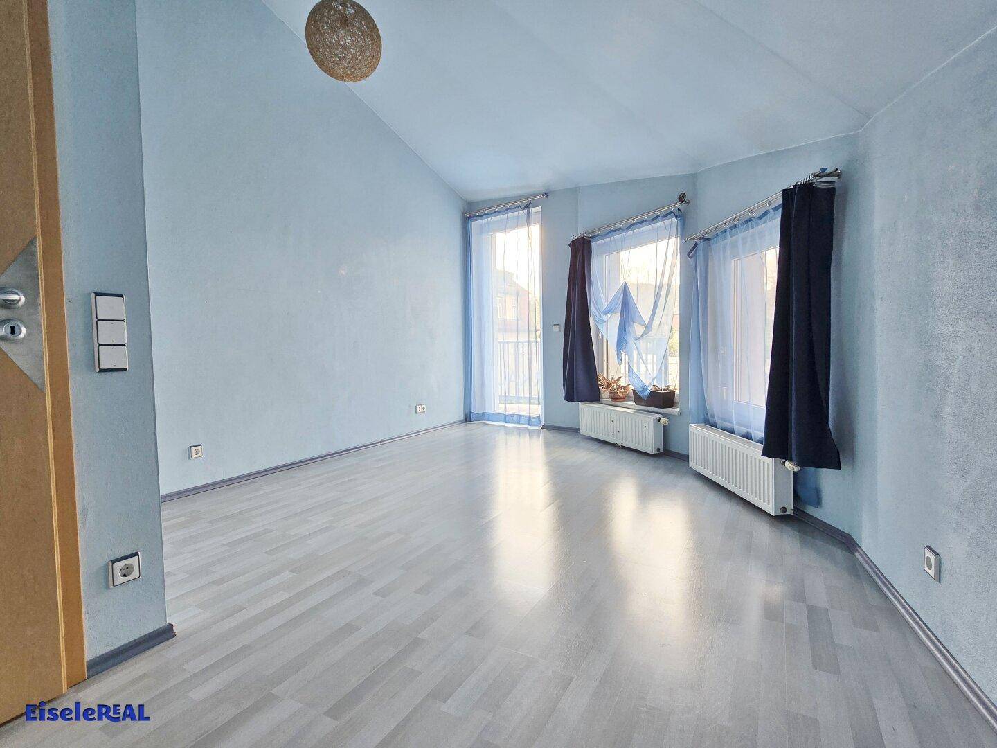 Helles Zimmer mit grauem Laminatboden, blauen Wänden und Zugang zu einem französischen Balkon.