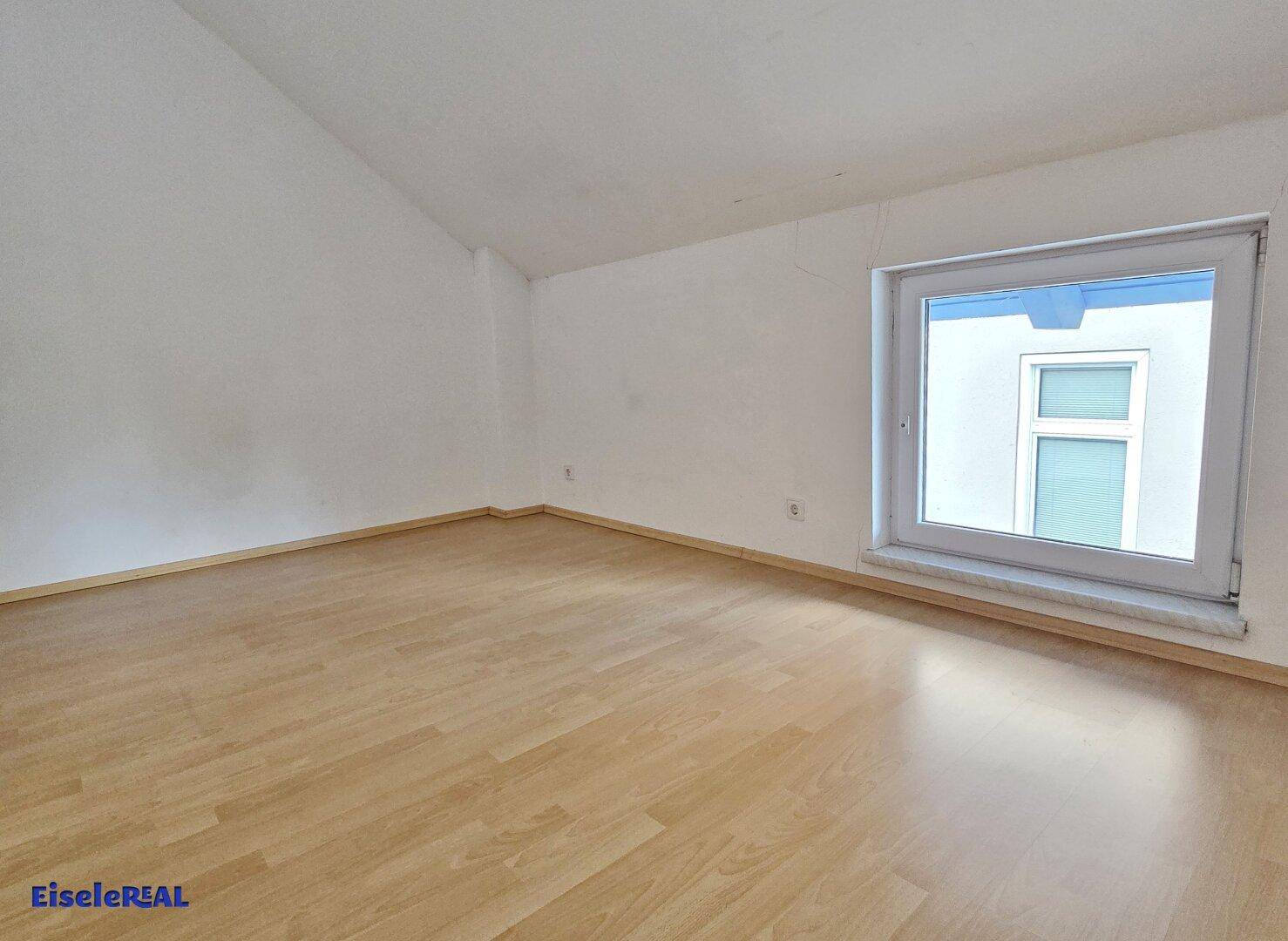 Leeres Zimmer mit hellem Laminatboden und einem großen Fenster, das viel Tageslicht hereinlässt.