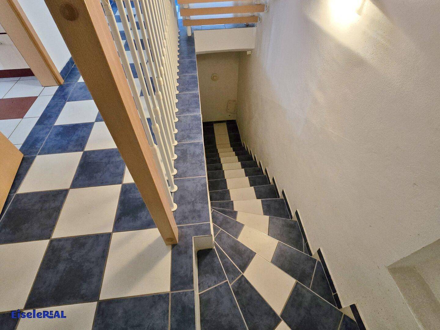 Treppenabgang in den Keller mit blau-weiß kariertem Fliesenboden und hellem Geländer.