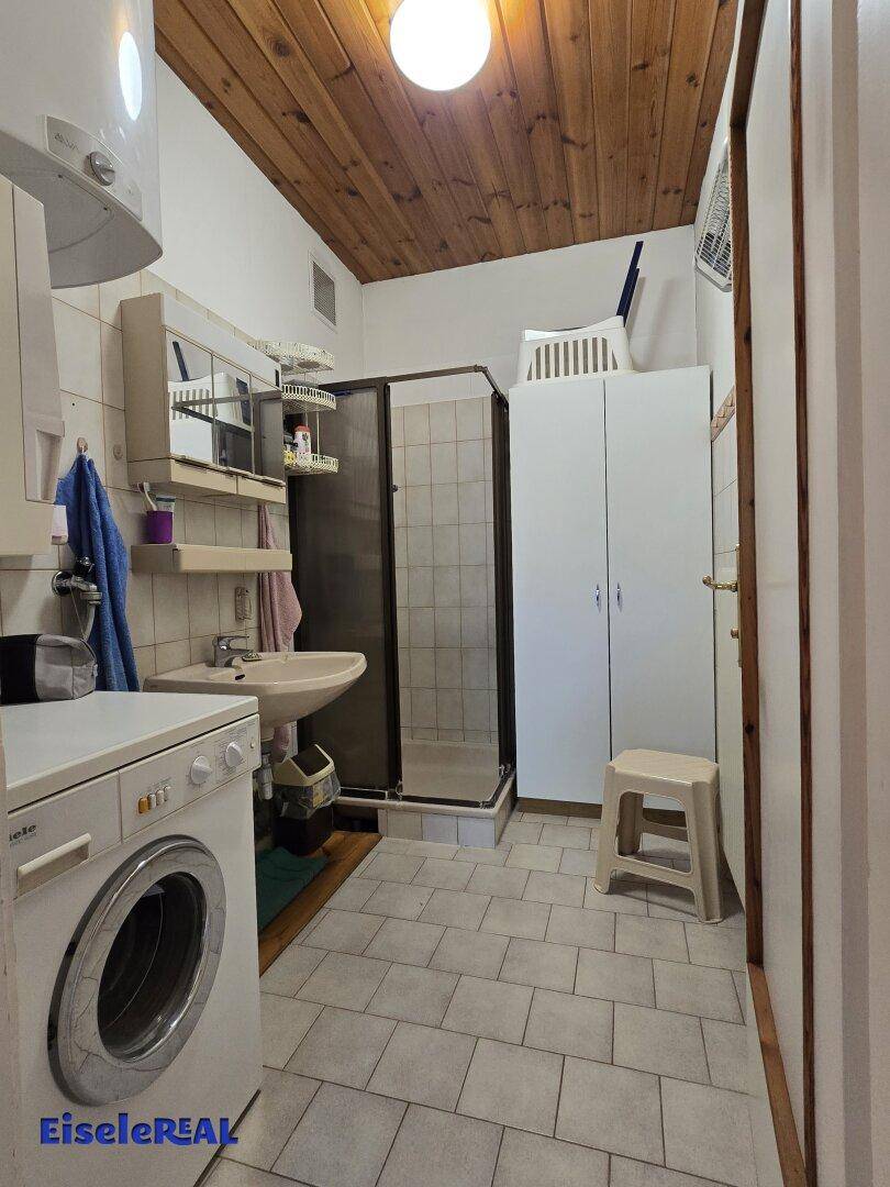 Badezimmer mit Dusche, Waschmaschine, Waschbecken und Holzdecke.
