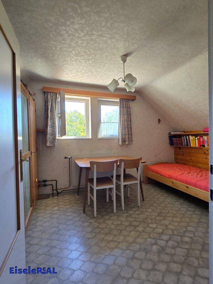 Dachzimmer mit schrägen Wänden, zwei Fenstern und einfacher Einrichtung.