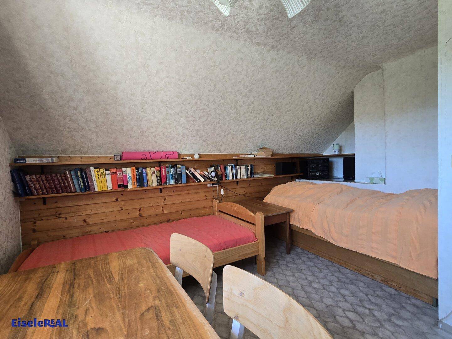 Dachzimmer mit zwei Betten, Bücherregal und schrägen Wänden.