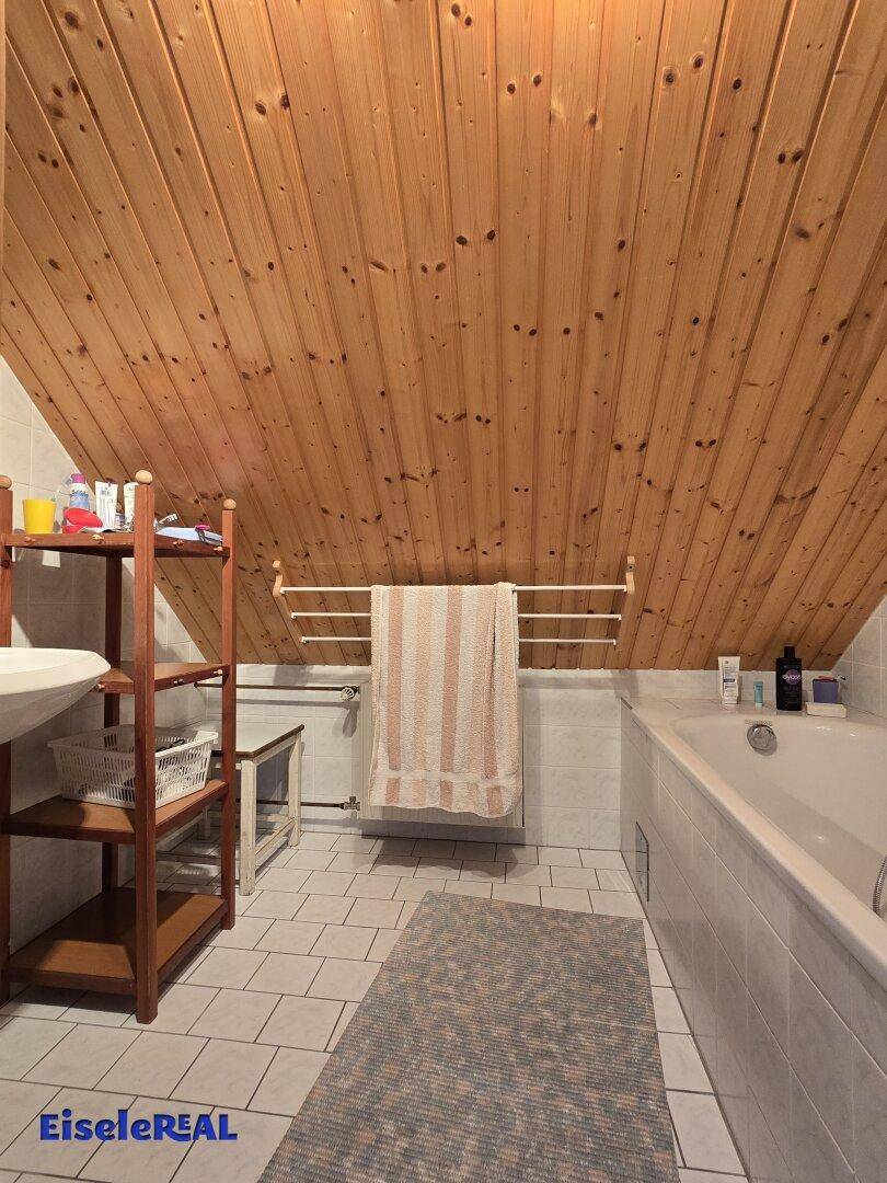 Badezimmer im Dachgeschoss mit Holzverkleidung an der Decke, Badewanne und hellen Fliesen.