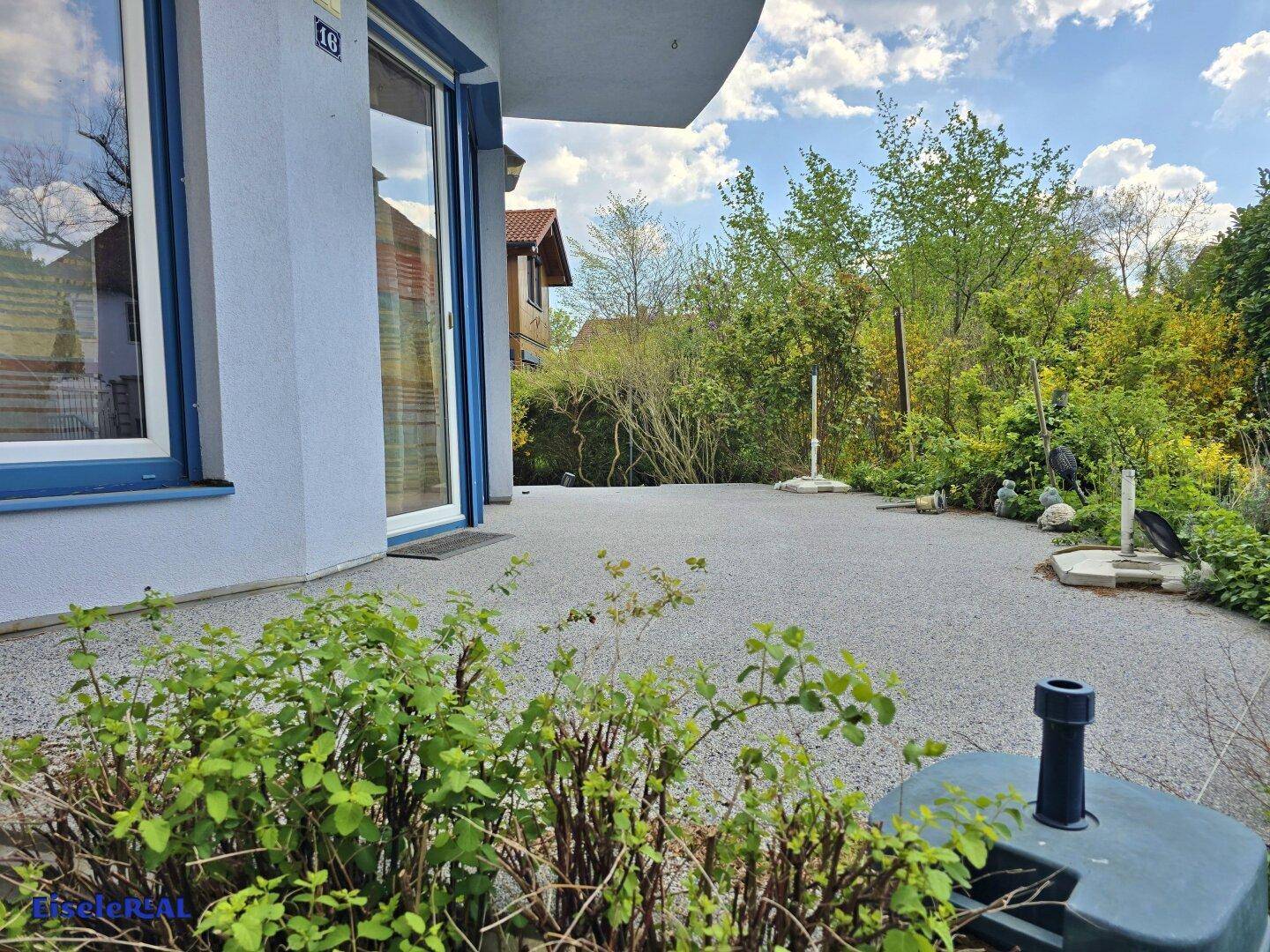 Eine gepflasterte Terrasse neben dem Haus, umgeben von grünen Sträuchern unter hellem Himmel.