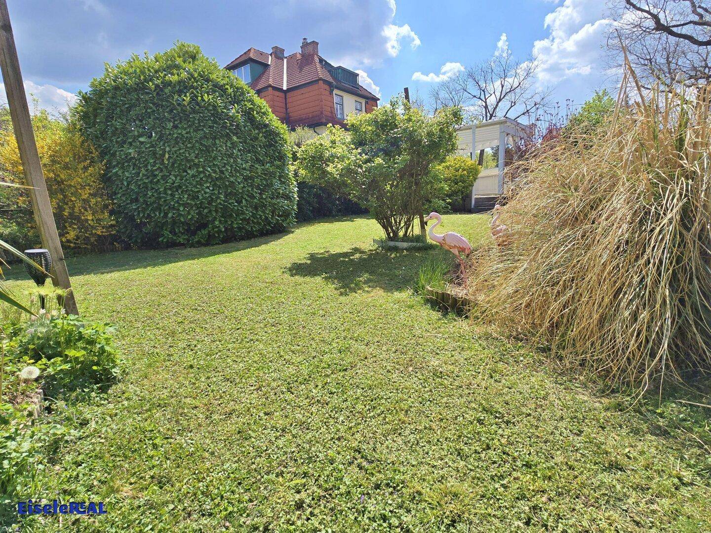 Ein gepflegter Garten mit Rasenfläche, hohen Gräsern und einem dekorativen rosa Flamingo.