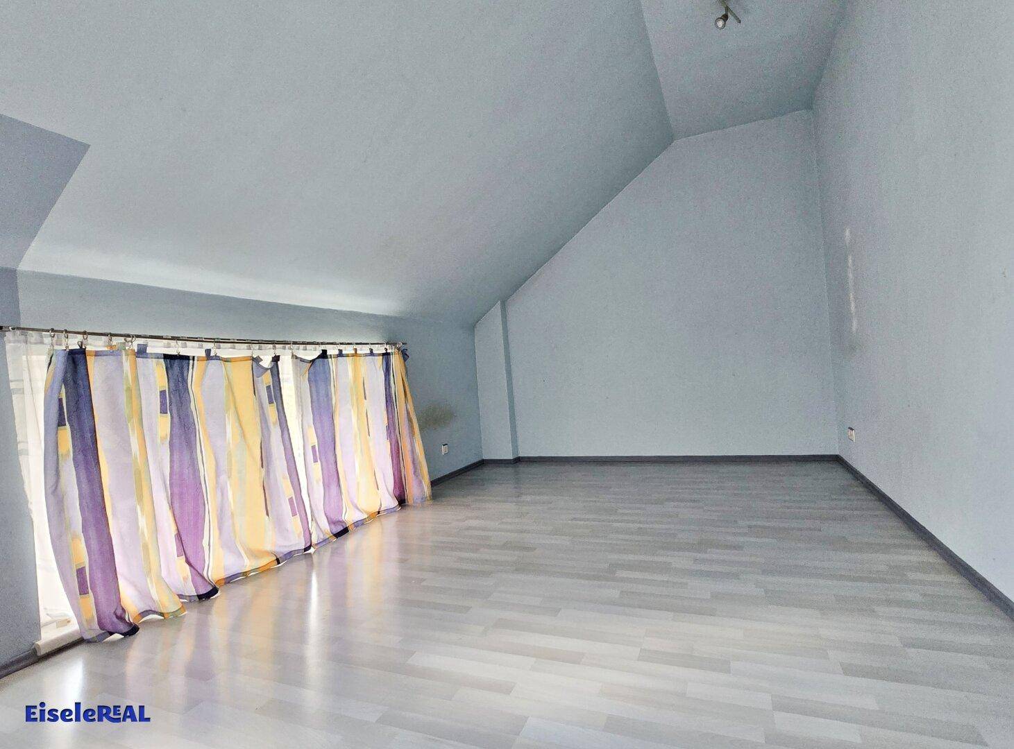 Dachzimmer mit schrägen Wänden, hellem Laminatboden und einem langen Fenster mit bunten Vorhängen.