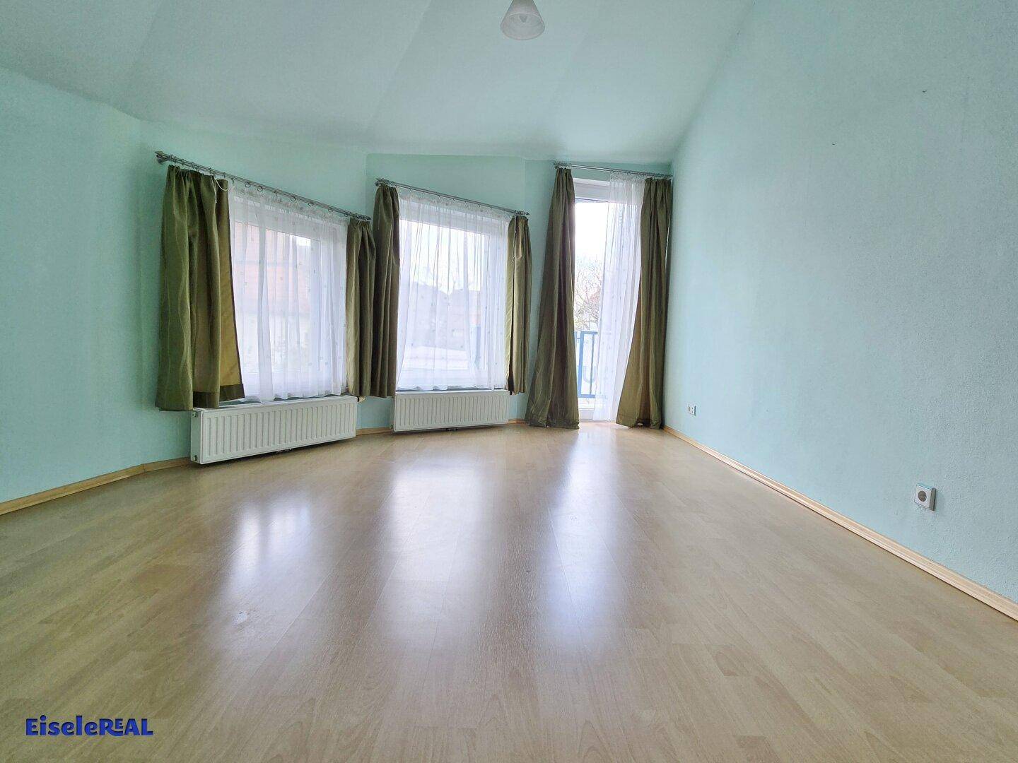 Heller Raum mit hellblau gestrichenen Wänden, Laminatboden und zwei Fenstern mit Vorhängen.