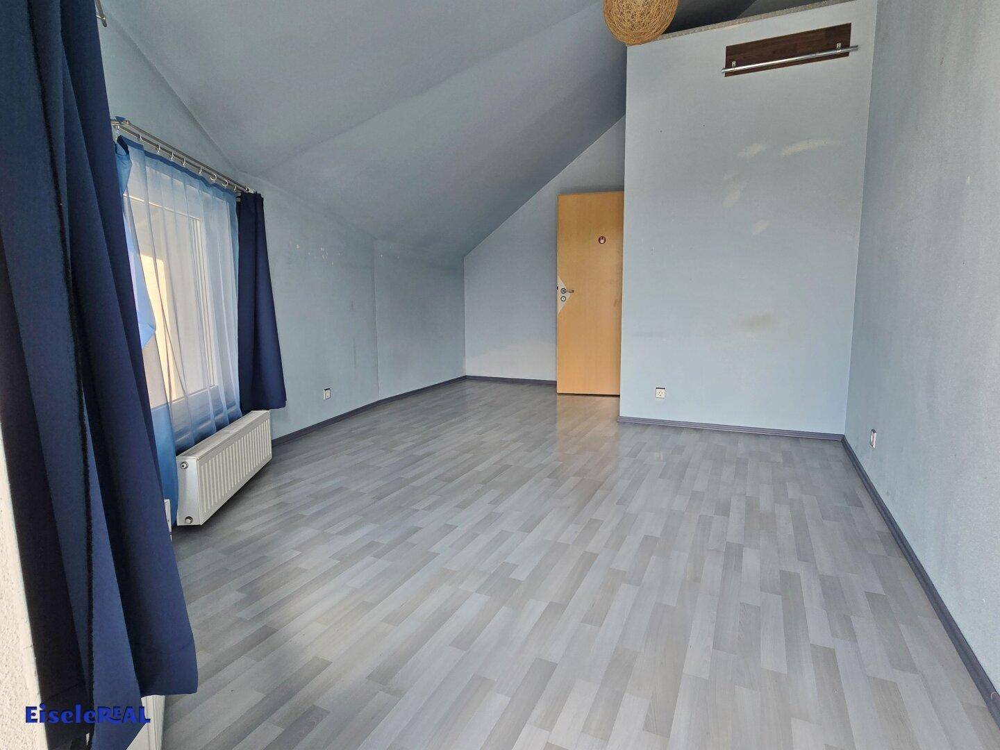 Dachzimmer mit hellgrauem Laminatboden, hellblauen Wänden und einem Fenster mit blauen Vorhängen.