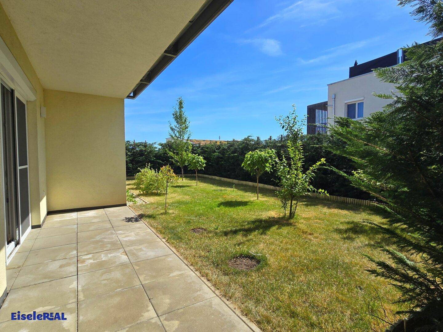 Geräumige Terrasse mit Fliesenboden und direktem Zugang zum grünen Gartenbereich unter strahlend blauem Himmel.
