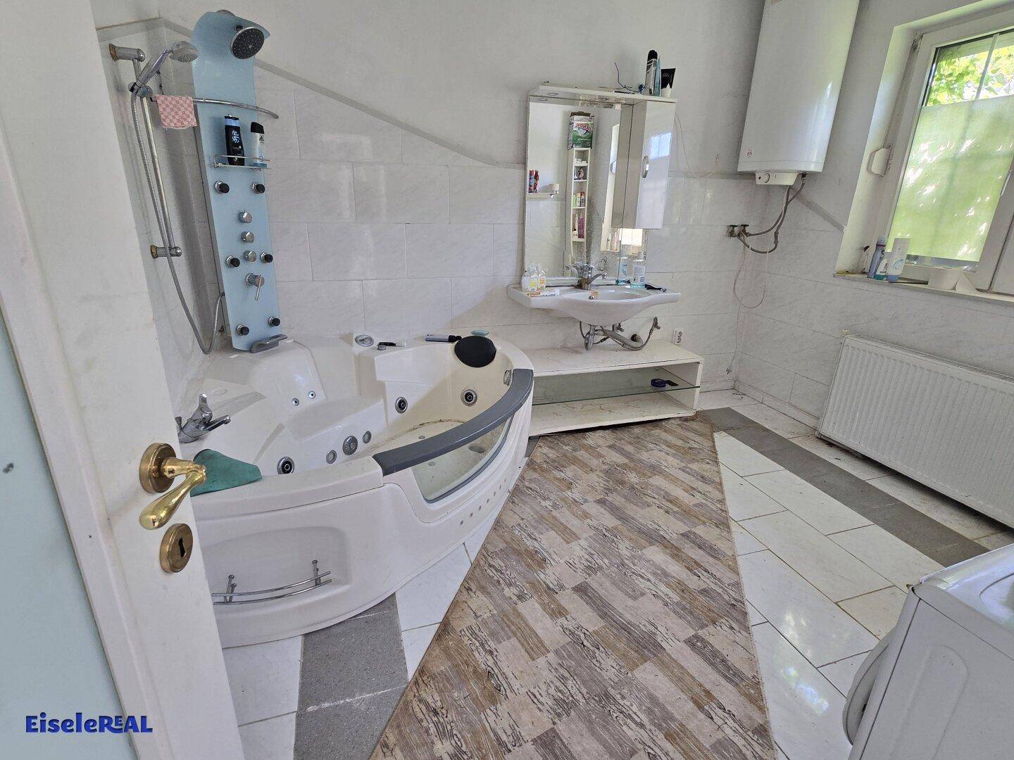Badezimmer mit Eck-Whirlpool, Waschbecken und Fenster, gefliesten Wänden.