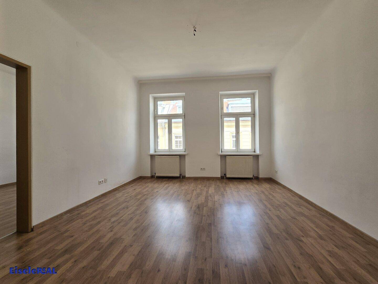 Geräumiges Wohnzimmer mit Holzfußboden und zwei großen Fenstern, die den Raum mit natürlichem Licht füllen.