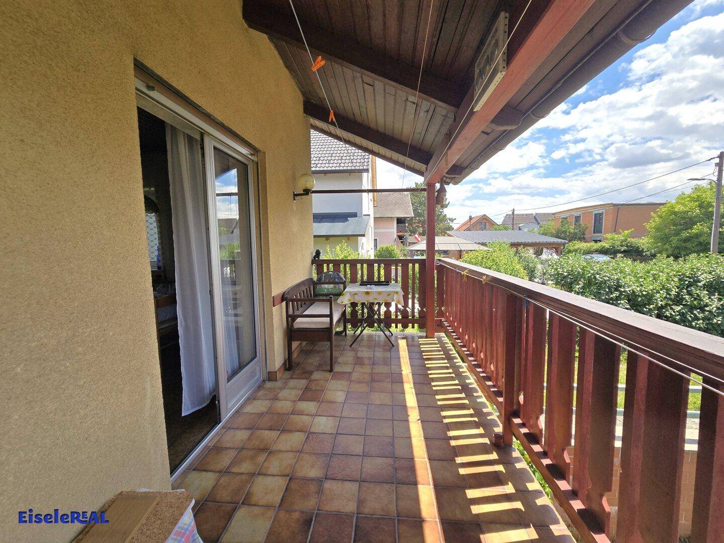 Überdachter Balkon mit Fliesenboden, Holzgeländer und einfacher Sitzgelegenheit im Freien.