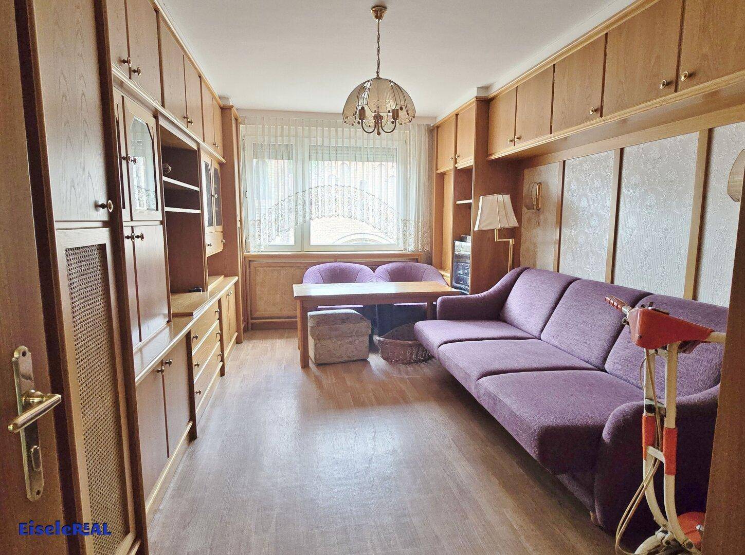 Gemütliches Wohnzimmer mit Holzvertäfelung, lila Sofa und einem kleinen Esstisch am Fenster.