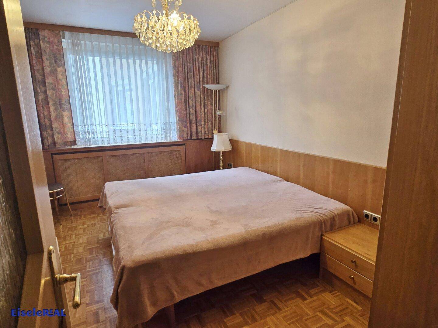 Schlafzimmer mit Doppelbett, Parkettboden und einem Fenster mit gemusterten Vorhängen.