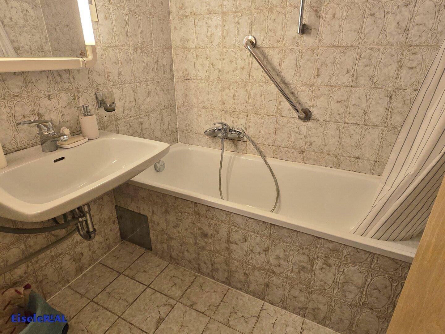 Badezimmer mit Badewanne, Waschbecken und gemusterten Fliesen, ausgestattet mit Haltegriffen.