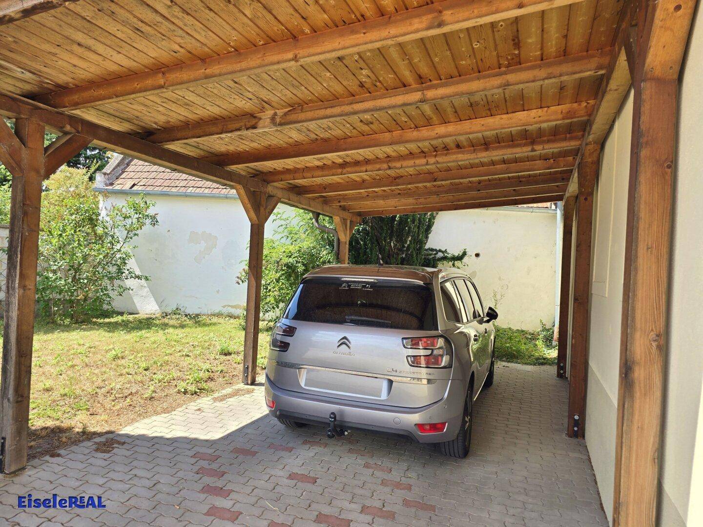 Überdachter Stellplatz für ein Fahrzeug mit Holzdach und gepflastertem Boden.