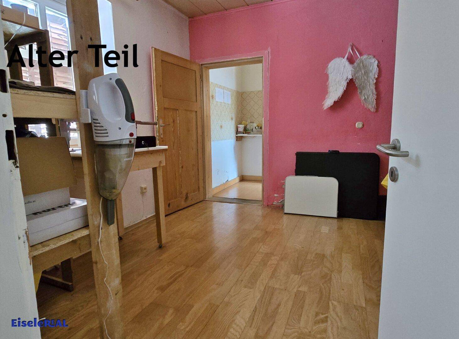 Einfacher Raum mit Holzfußboden und rosa Wand, der zu einem weiteren Bereich führt.