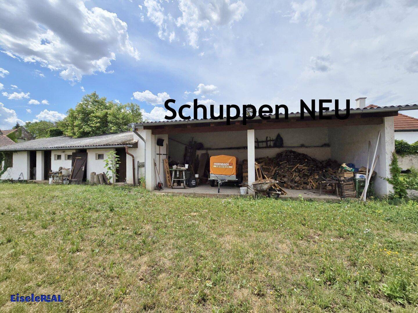 Offener Schuppen mit Holzlager und Grasfläche davor, nützlich für Fahrzeuge oder Gerätschaften.